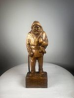 Front view of the hand-carved wooden figure showing detailed facial features, hat, coat, staff, and base.