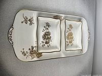Photo showing two rectangular bone china serving trays with gold chrysanthemum flowers and gold gilt edges and handles, placed nested inside each other on a gray surface.