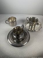 Three silver coffee cups and two saucers including a silver-plated serving tray. Cups vary in metal type and markings. Saucers have ornate edges.