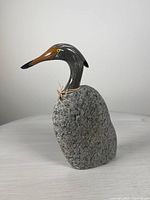 Side view showing the polychrome wooden head and natural gray stone body of the duck sculpture with raffia tie.