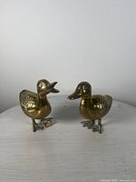 Two vintage solid brass duck figurines placed on a light surface against a white background. Both ducks are upright, one with an open beak and the other with a closed beak, showing overall condition and design.