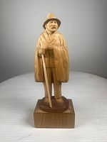 Front view of the hand-carved wooden traveler figurine showing detailed facial features, hat, cloak, and staff, mounted on a wooden podium.