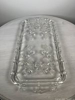 Top-down view of rectangular clear crystal serving plate with embossed roses and frosted leaf pattern beneath surface. Plate has handle-like edges and beaded border detail.