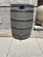 Front view of brown and black plastic rain barrel with metal spigot positioned on concrete patio against a wall.