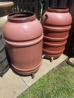 Two terracotta plastic water barrels standing upright outside on pavement next to garden soil. Both barrels are open top, showing screw threads. Both have ribbed reinforcement rings around middle and bottom. One barrel is more rounded smooth and the other features horizontal ribs.