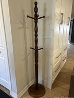 Full view of the entire wooden coat rack standing in a corner beside white cabinetry, showing its tall height and round base.