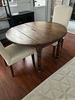 Oval antique wooden table with dark wood finish shown with both drop leaves open, positioned between two beige upholstered chairs on wood floor.