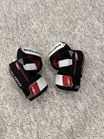 Pair of Bauer black and red youth elbow pads laid on carpet, showing overall front and top view.