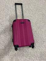 Front view of a dark pink hardshell carry-on suitcase with the retractable handle extended, positioned on a carpeted floor.