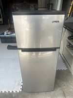 Front view of stainless steel Magic Chef mini fridge with freezer compartment on top, standing in a garage space.