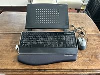 Top-down view of Microsoft black wireless keyboard with ergonomic wrist rest, wired mouse, and black laptop stand on wooden table.