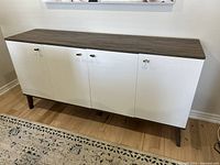 Front view of the sideboard showing wood finish top with four white doors and metal handles, standing on wood finish tapered legs.