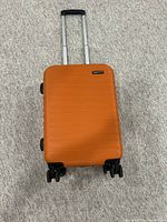 Closed orange hard-shell carry-on suitcase with four spinner wheels and telescopic handle retracted, placed on a carpeted floor.