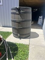 Full side view of the brown plastic rain barrel with black bands, placed on concrete outdoors.
