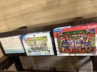 Three puzzle boxes displayed on a wooden floor showing GrayMalin beach puzzle and two Ravensburger puzzles including cottage garden and Christmas holiday themes.