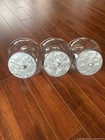 Three transparent round glass jars with silver metal lids placed on a wooden floor, view from metal lid side showing textured metal finish.