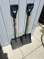 Two hardwood-handled D-handle shovels leaning against a wall, showing blade size and handle style