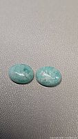 Two oval amazonite cabochons on a textured gray surface showing smooth polished blue-green stones with natural inclusions.