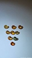Top-down photo showing a collection of 10 small round faceted sapphires of various colors including yellow, orange, and green on a light surface.