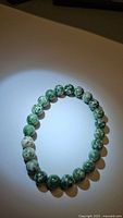 Photo of a stretchy bracelet made of round, 8mm spotted jasper beads with green and beige natural patterns laid flat on a light surface under light focused illumination.