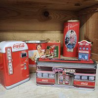 Multiple Coca Cola collectible items displayed on wood shelf, including ceramic candle holder, metal tins shaped like vending machines and diners in red, white, and blue colors.