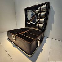 Opened brown leather-like case with six metal shot glasses attached to lid and compartmentalized interior with straps.