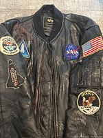 Front view of worn black leather jacket showing multiple NASA and space mission patches, US flag on sleeve, ribbed collar and zipper front.