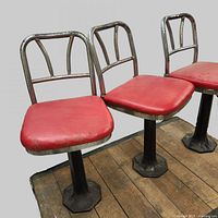 Three vintage soda fountain/diner stools arranged on wooden flooring, showing red vinyl seats and chrome frames with cast iron bases.