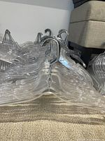 Multiple stacks of clear plastic clothes hangers with metal hooks lying on carpet and stacked against a wall.