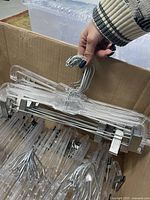 Box filled with transparent plastic pant hangers with metal clips and hooks, showing several hangers stacked inside the box with a person holding a hanger.