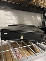Front view of the closed black Star metal cash box on storage shelf, showing locking key inserted.