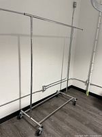 Full view of metal clothing rack against wall, showing horizontal hanging bar and caster wheels.