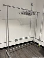 Full view of metal clothing rack with clear plastic hangers on crossbar, placed against light-colored wall with visible electrical piping.