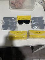 All seven pairs of sunglasses individually wrapped, displayed with yellow branded cases on white table.