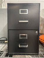 Front view of black 2-drawer filing cabinet showing slight dent on top drawer, silver handles and label holders, bottom drawer with lock.
