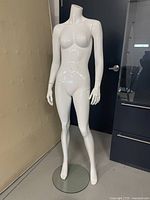 White headless full body female mannequin standing on round clear glass base, positioned near a door indoors.