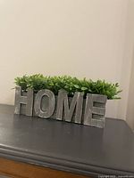 Decorative HOME planter with faux greenery placed on a dark surface against a plain wall