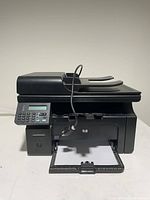 Front view of black HP LaserJet multifunction printer showing paper input and output trays with power cable.