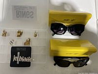 Two pairs of shiny black sunglasses in bright yellow Shady Lady cases, an open jewelry organizer with various gold necklaces, a padlock pendant, and a jeweled black hairpin on card.