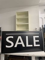 Front view of black SALE sign with white lettering, held upright against a white wall with shelving in background.