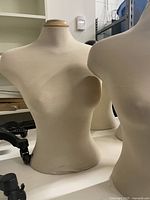 Two light beige fabric covered half body female mannequins with wood-colored tops, viewed from the front and side, placed on white surface in an indoor storage or office environment.