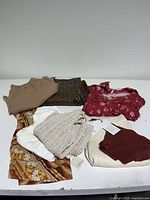 Seven clothing items laid out flat showing variety of styles, colors, and textures including knit vest, floral tops, ribbed shorts, and printed pants in earthy tones.