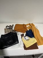 Photo showing assortment of folded clothing including black faux leather pants, white pants, checked and knitted tops in mustard yellow and brown.