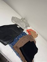 Eight clothing items laid flat including light patterned top, black pants, denim shorts, beige and brown shorts, black ribbed bike shorts, and various tops and bralettes.