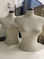 Two fabric-covered half-body mannequins with black spherical tops shown side-by-side on a white surface in a room with shelving and a small wooden cupboard in the background.