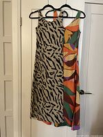 Two dresses hanging on black plastic hangers on a door. One dress is beige with black tiger stripes, the other has a colorful abstract pattern in green, orange, purple, and yellow.