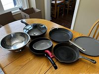 Top view of all cookware items arranged on wooden dining table including colander, pans with lids and handles, pizza pan.