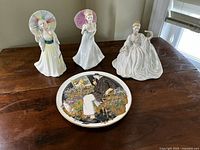 Overall photo showing three Royal Doulton figurines and one decorative plate on wooden table.