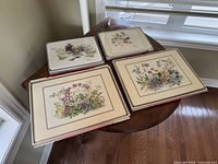 Four placemats laid out, showing detailed prints of flowers, berries, and birds on cream backgrounds with brown borders.