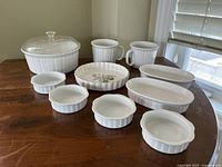 Wide view of full set of 10 vintage Corning Ware dishes arranged on wooden table including casserole with glass lid, mugs, oval baking dishes, and small ramekins.
